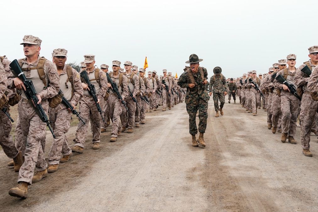 Marine Corps recruits march down a road on two sides while a drill instructor shouts from the middle of the dirt road during an overcast day.