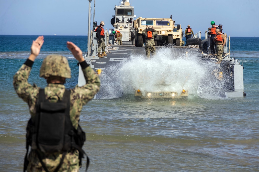 A Marine signals to a transport vessel landing ashore offloading a military vehicle during the day.