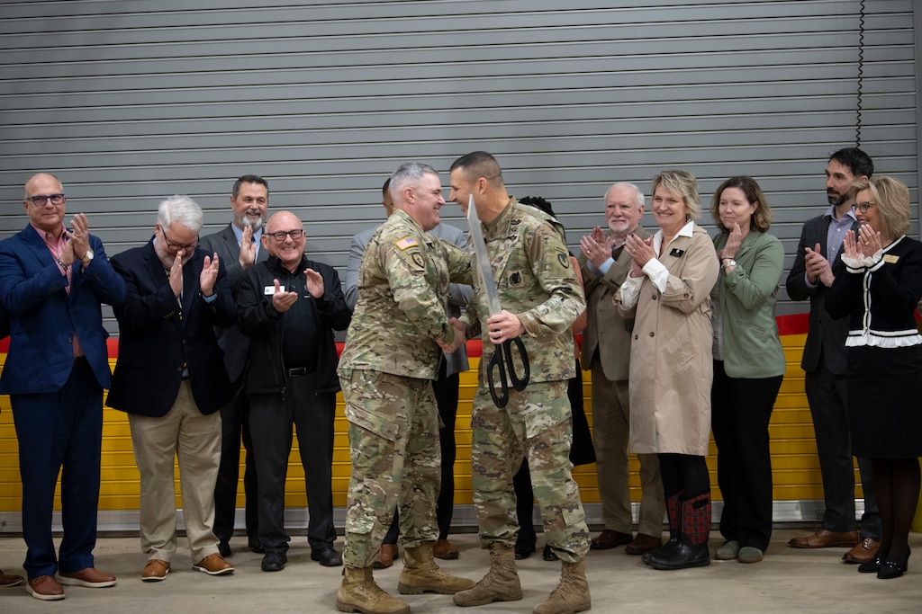Two Army officers in camouflage uniforms shake hands with people clapping in the background.
