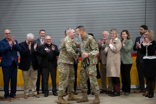 Two Army officers in camouflage uniforms shake hands with people clapping in the background.