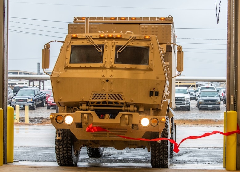Beige military vehicle breaks a red ribbon as it enters the vehicle maintenance facility at Red River Army Depot.