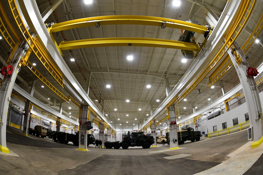 Interior view of a large warehouse equipped with yellow overhead cranes with military vehicles in the background.