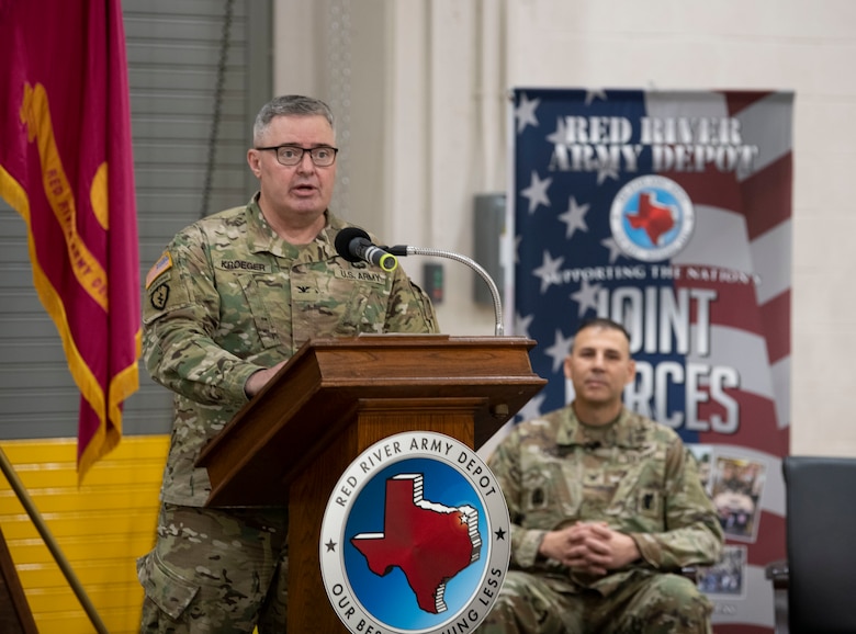 Col. Calvin Kroeger speaking at a podium with Col. Denis Fajardo sitting in the background.