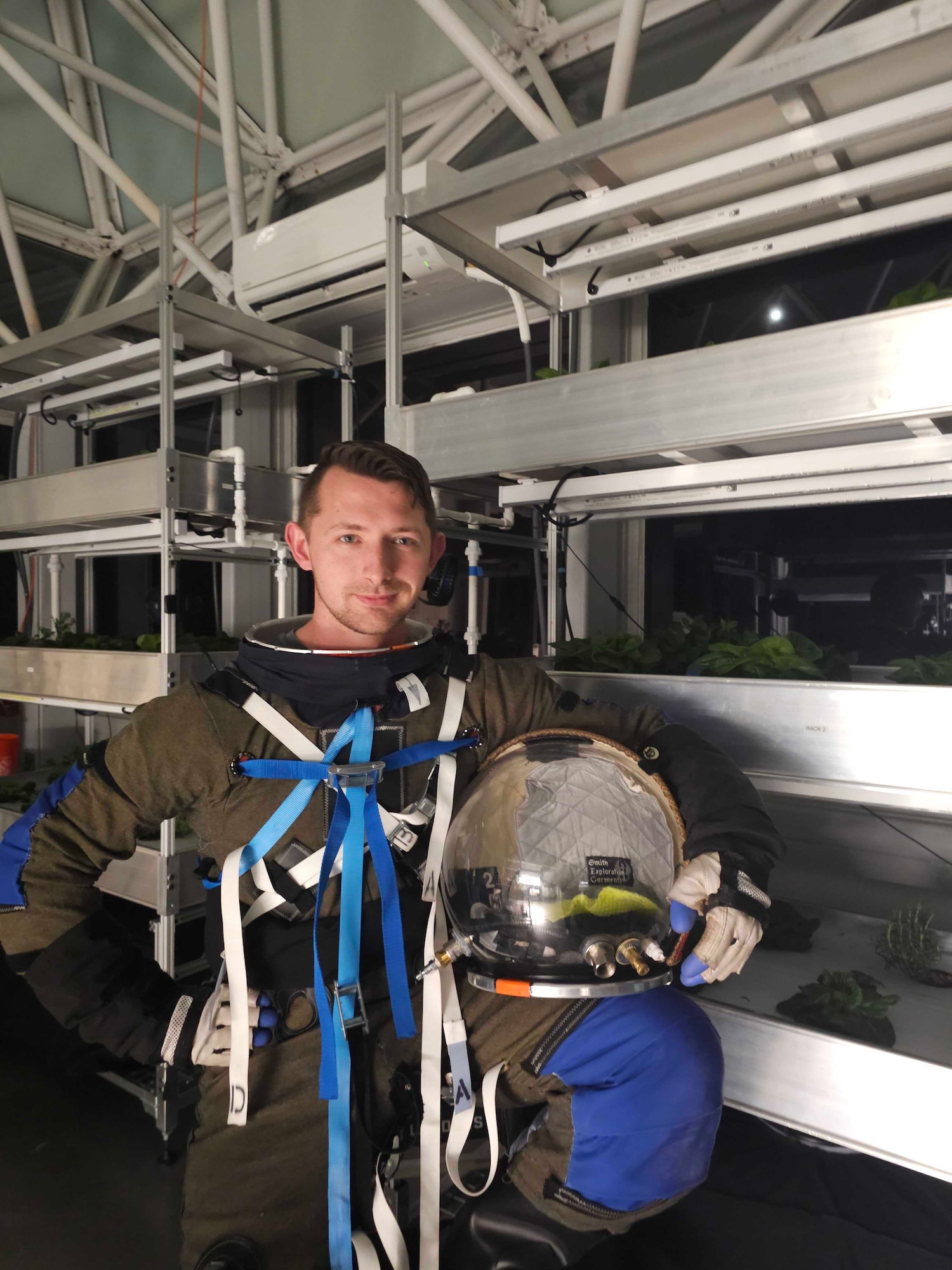 Wallace stands next to vegetation racks while he wears a space suit used in the study