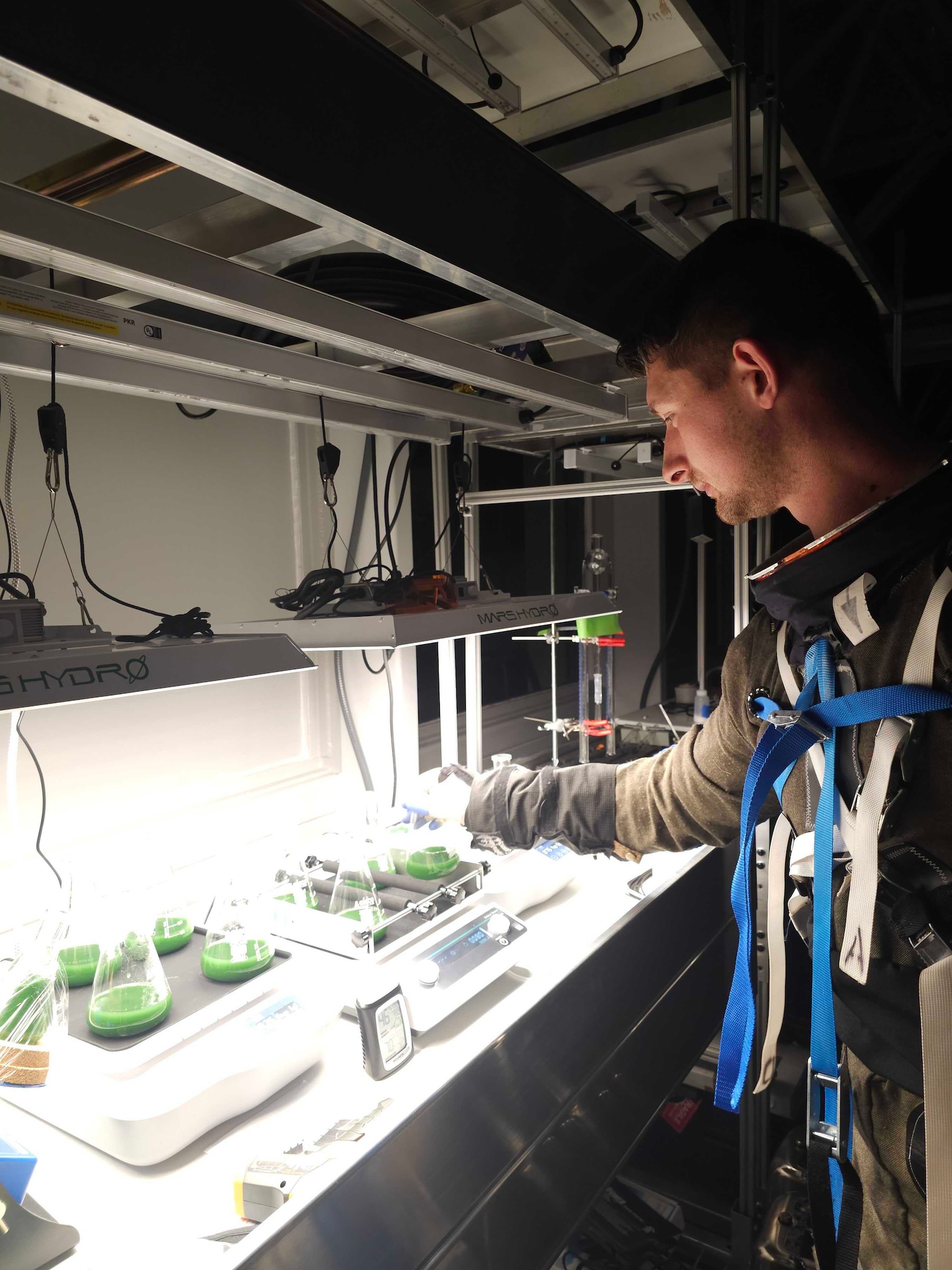 Wallace is standing next to a lab table that has on it multiple lab equipment and flask filled with algae.