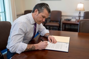 A man sits at a desk inside an office signing a document with his right hand.