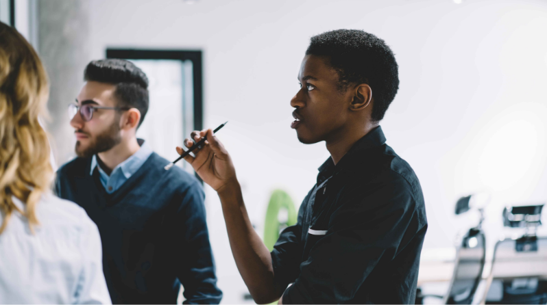 Two men and one woman stand together in front of a whiteboard. One man holds a pencil while contemplating the information.