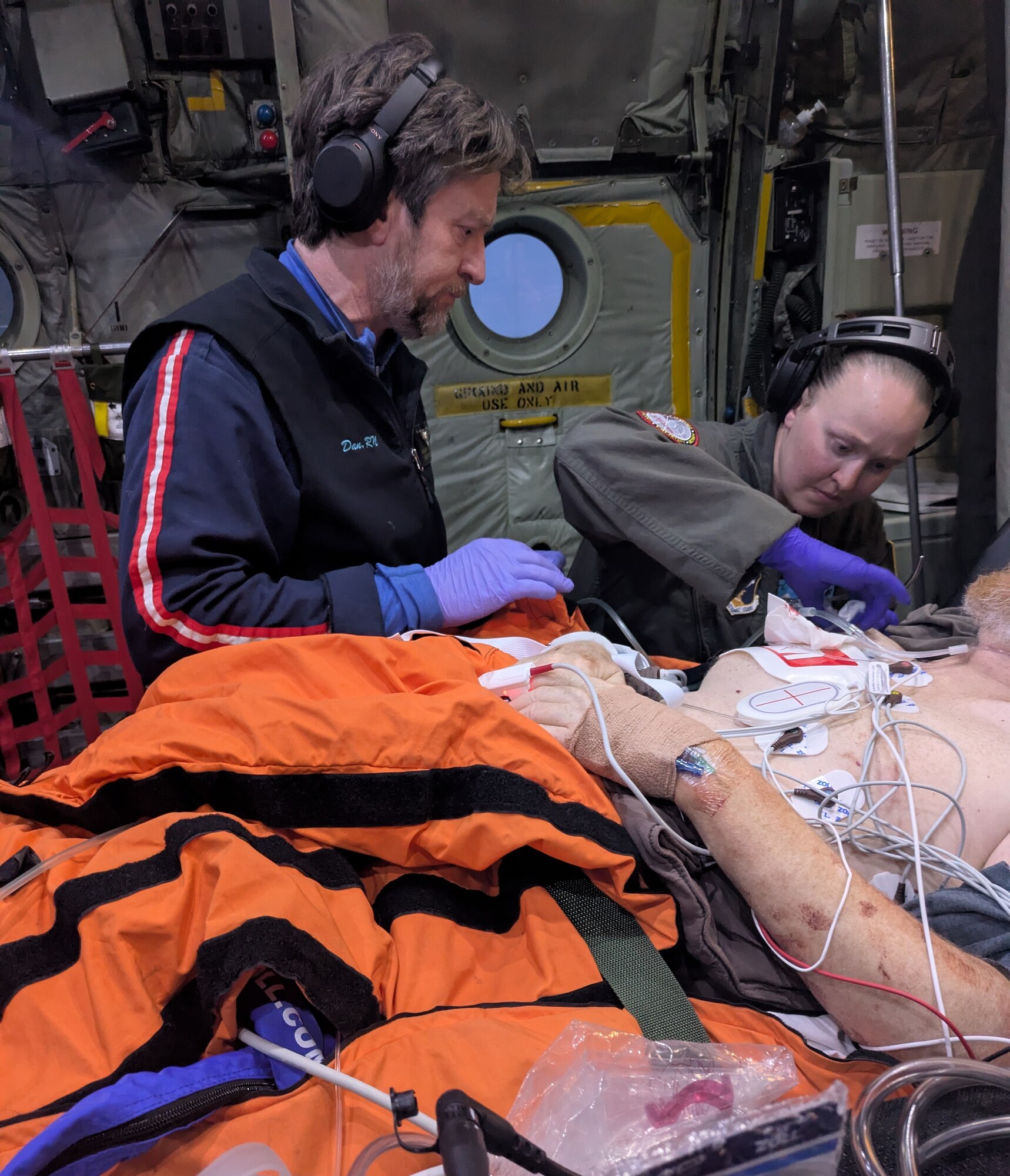 Two nurses apply medical care to a patient on a gurney in the cargo hold of a airplane