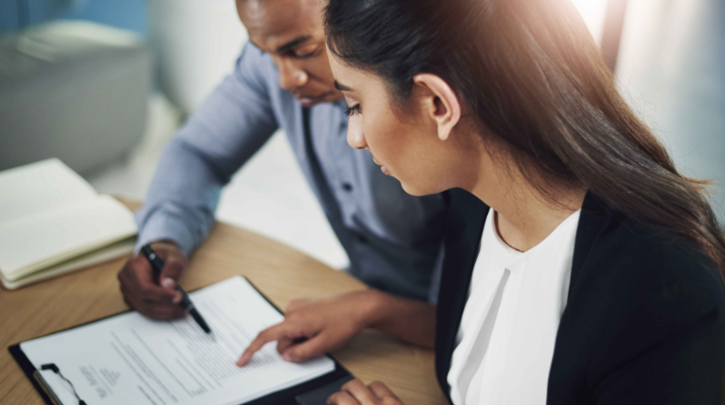 A man and a woman in business attire sit at a table, reviewing paperwork on a clipboard.