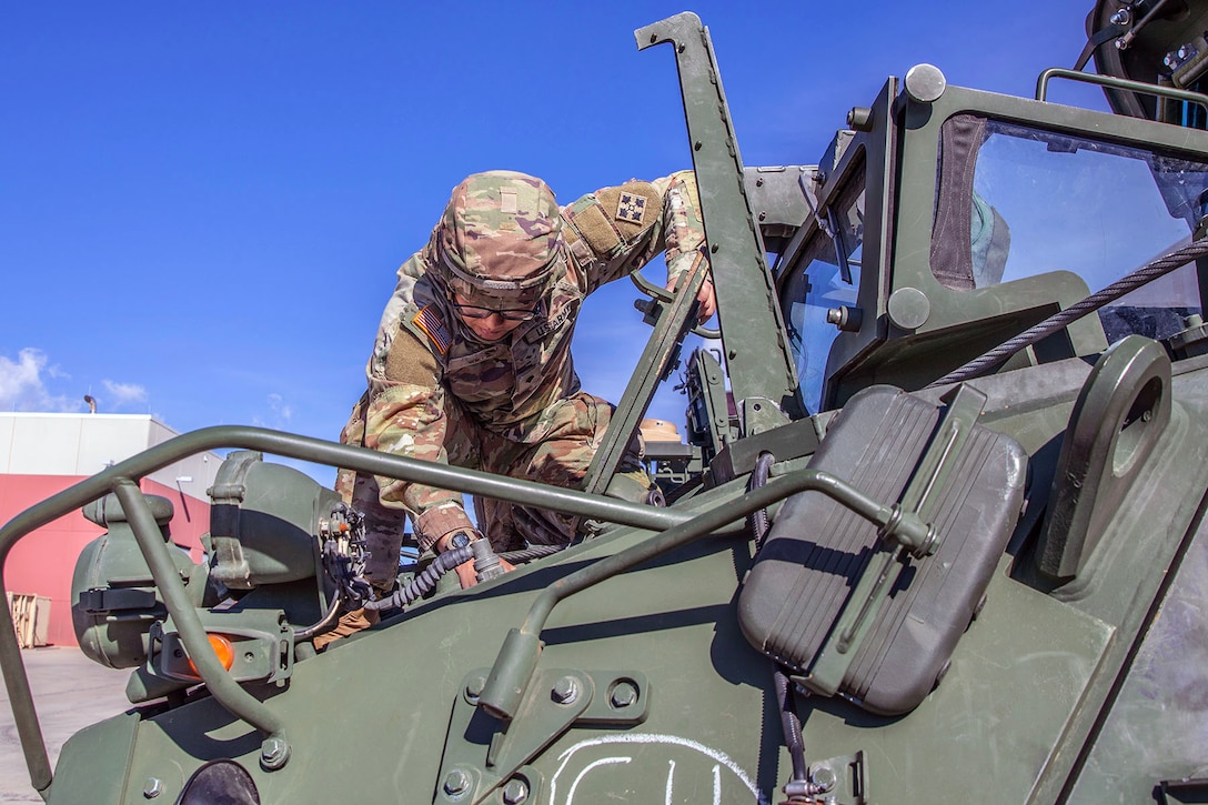 A soldier in uniform works on a green armored vehicle under a blue sky.