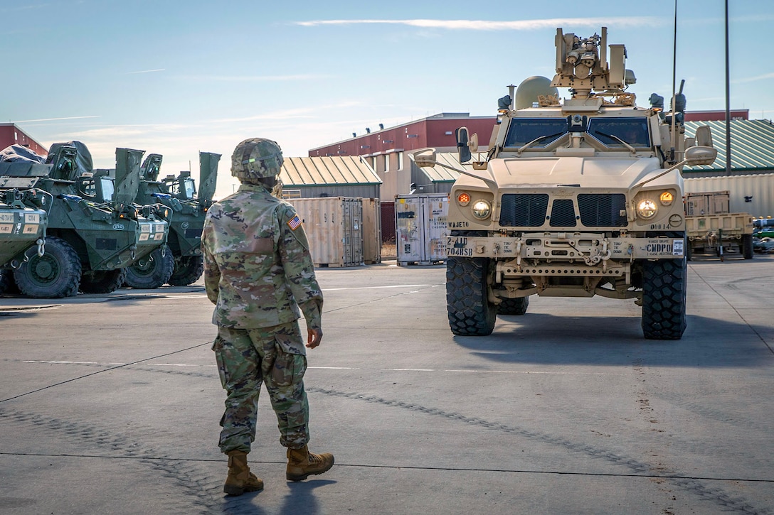 A soldier directs a vehicle as other vehicles sit to the left under a blue, cloudy sky.