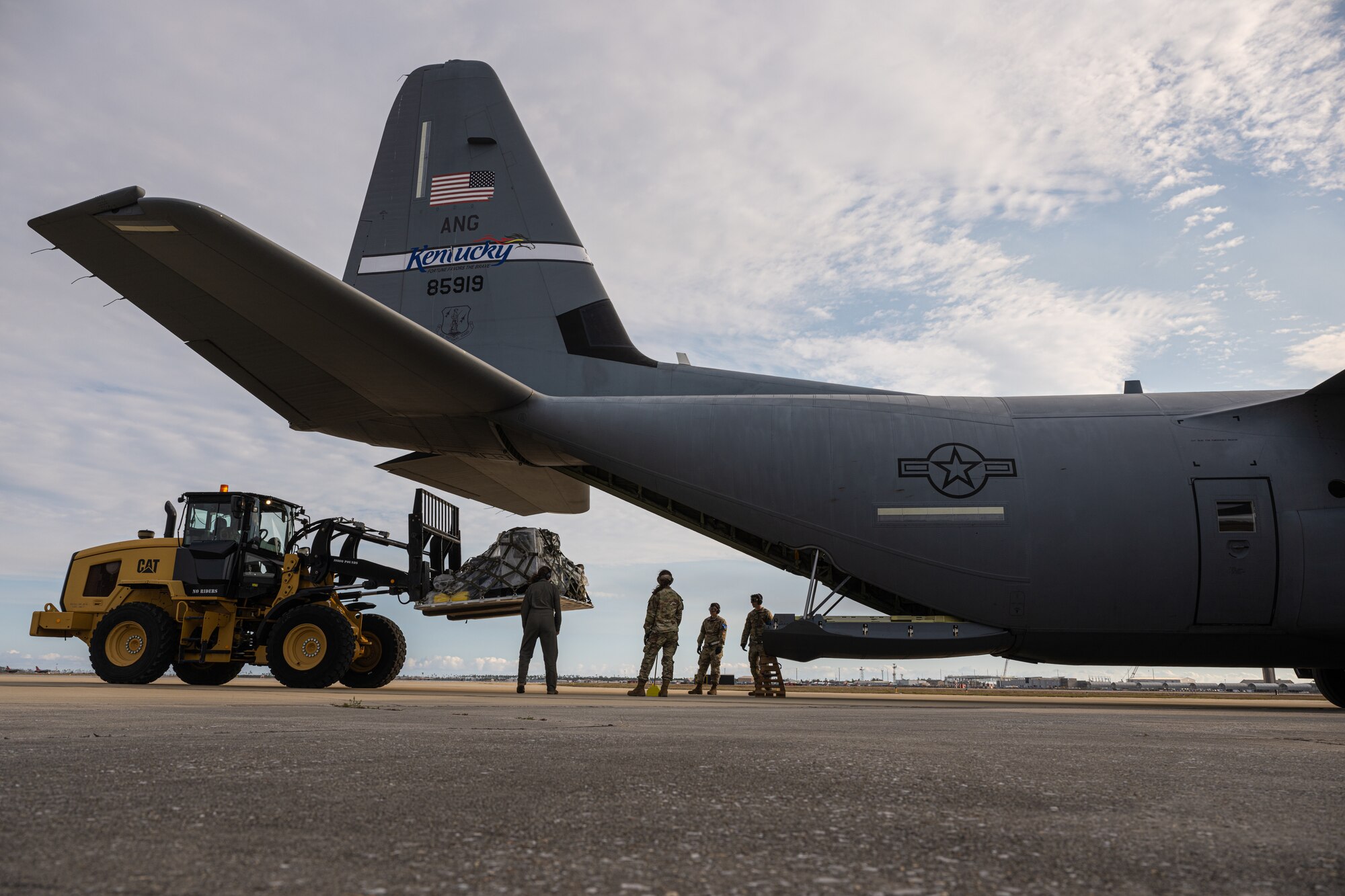 A photo of a crane lifting equipment into a C-130 jet with Airmen standing around.