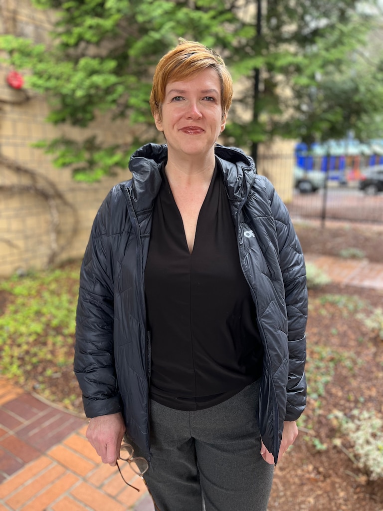 A woman standing with trees and a building behind her.