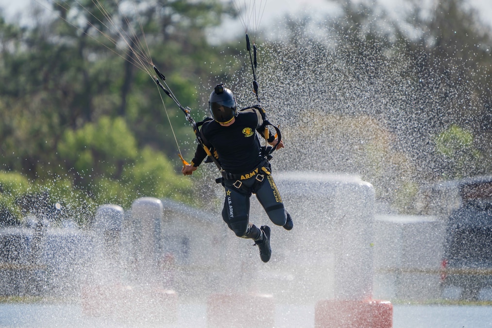 A soldier in black gear holds parachute strings while descending as water splashes upward.