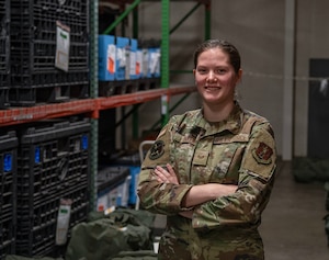 A female airman wearing green military uniform stands with arms crossed in large storage bay.