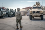 A soldier walks in front of an armored military vehicle during the day.