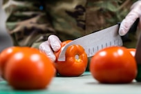 Closeup of a hand holding a knife slicing through a tomato with more tomatoes in the foreground.