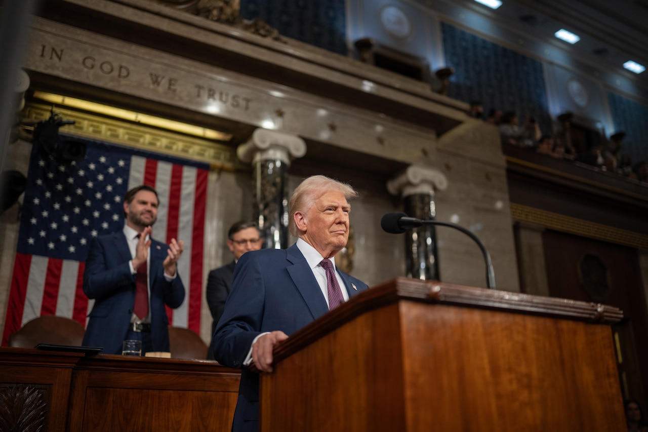 A man in a suit stands behind a lectern while two men applaud behind him.