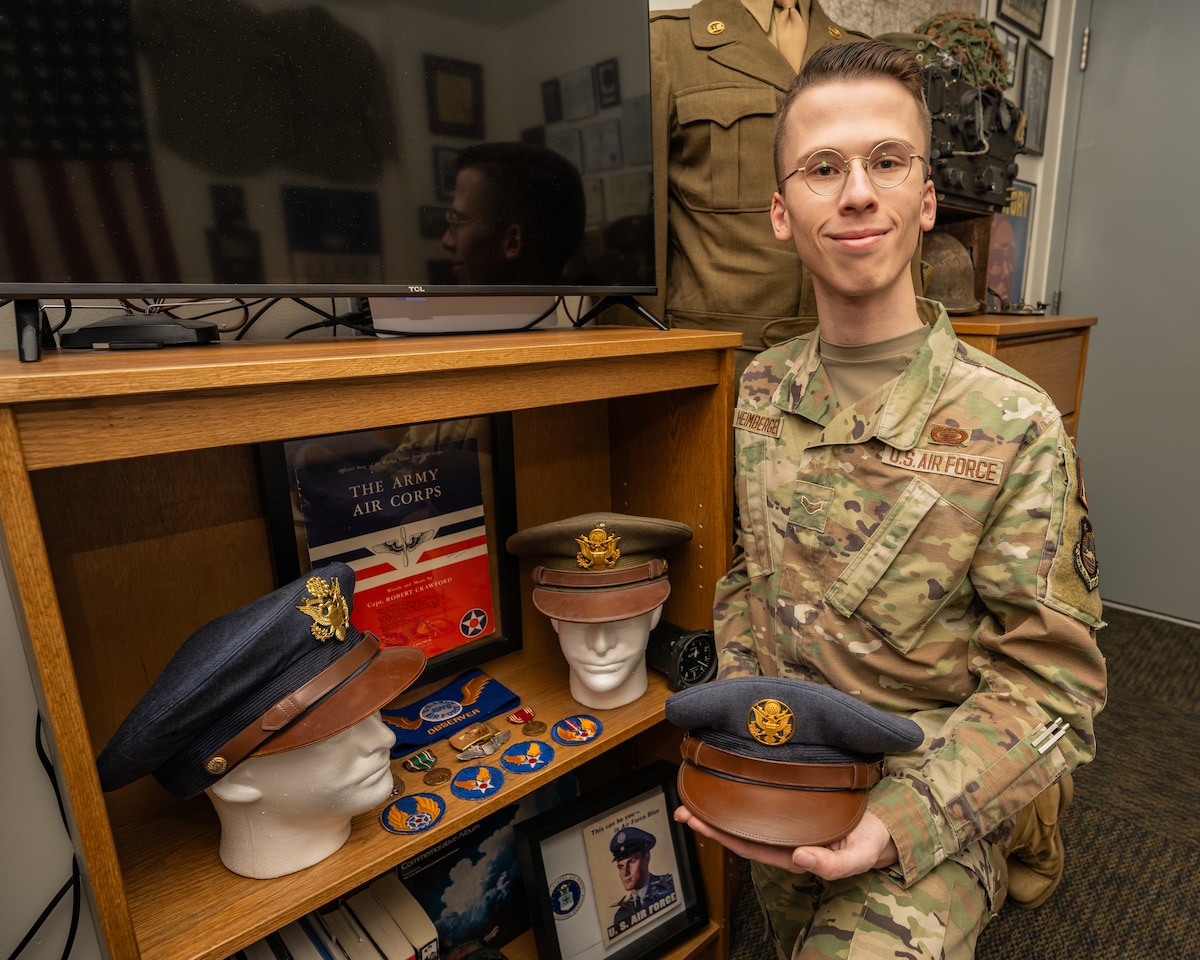 A young Airman in modern military camo uniform poses with a vintage Air Force service cap with brown leather visor