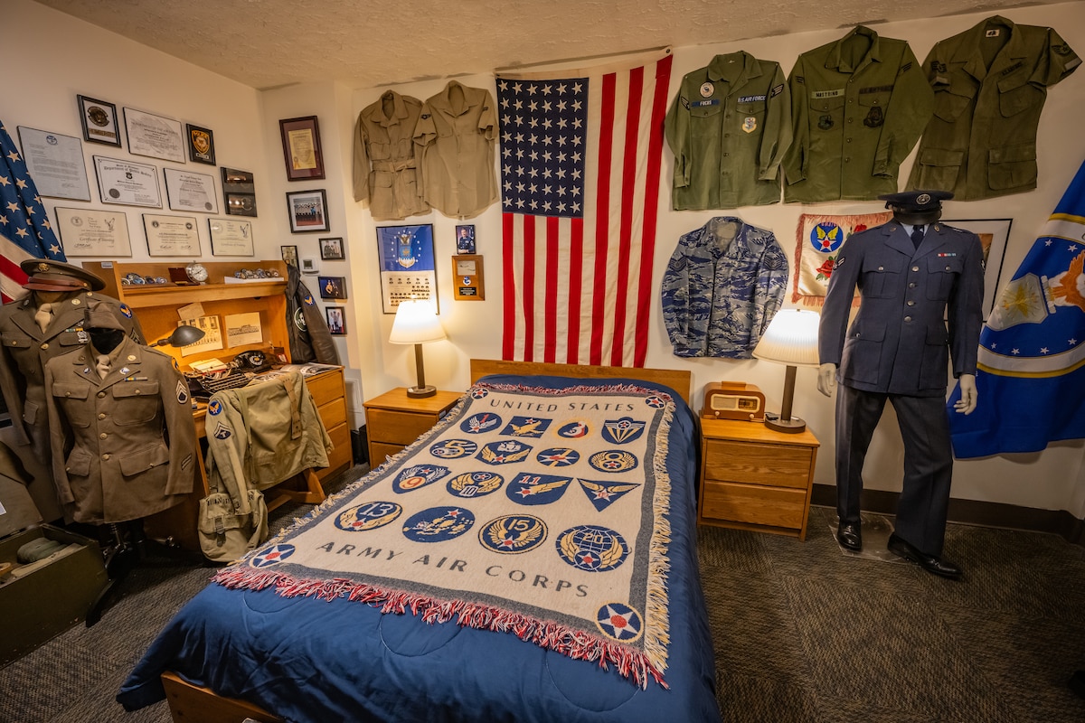 Vintage military uniforms and memorabilia on display, a U.S. flag hangs above the headboard of a bed with a throw blanket with the 16 original numbered Air Force patches displayed