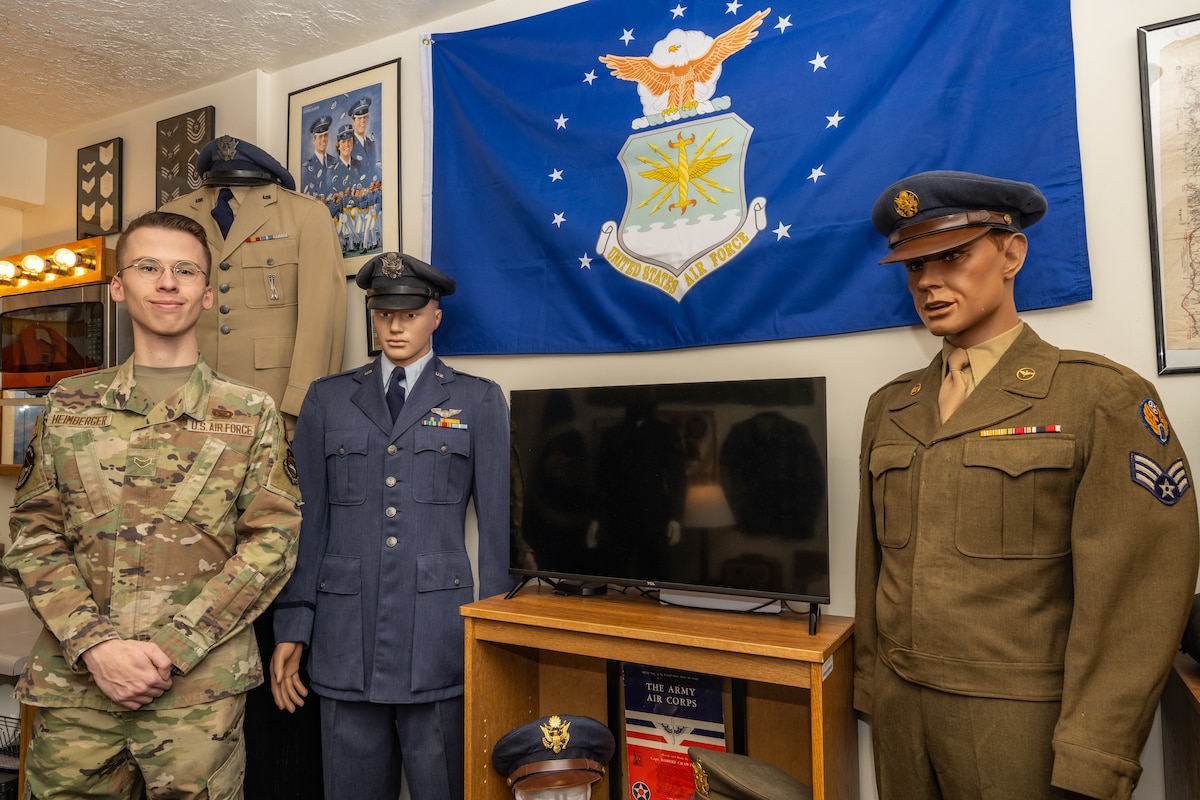 A1C Heimberger wears a modern military camo uniform, stands next to two mannequins wearing vintage military service dress uniforms, one blue one greenish-brown