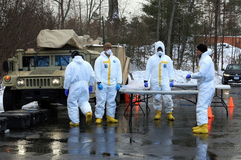 Personnel in white protective suits gather outdoors around a table in front of military equipment.