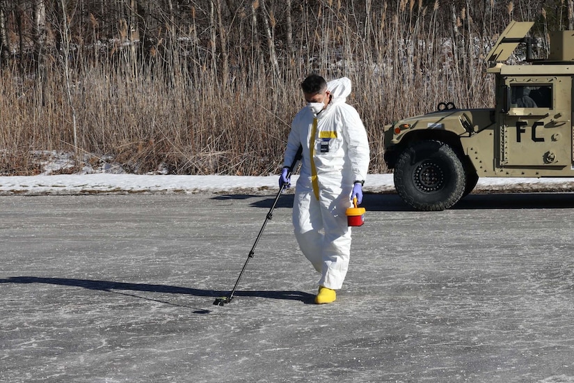 A man in a white protective suit walks across a parking lot, waving a long-handled equipment along the ground. He carries a small container in his left hand.