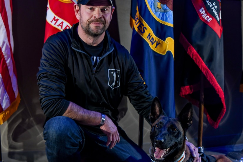 A man kneels next to a dog laying down in front of flags.