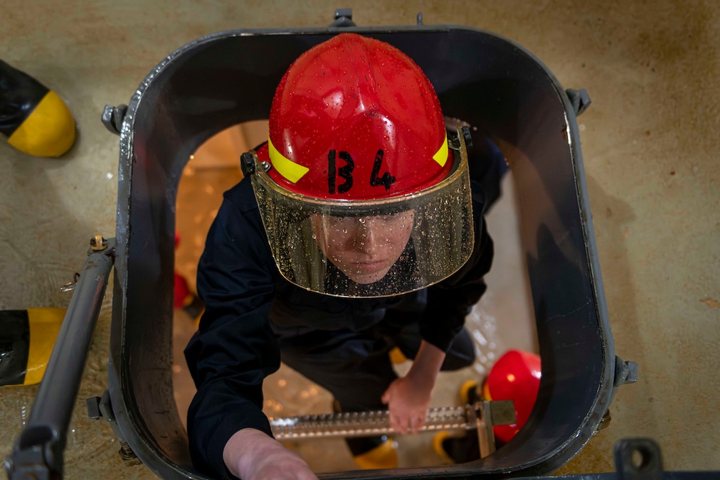 A sailor wearing a red helmet with a visor climbs a vertical ladder to another floor in a naval ship.