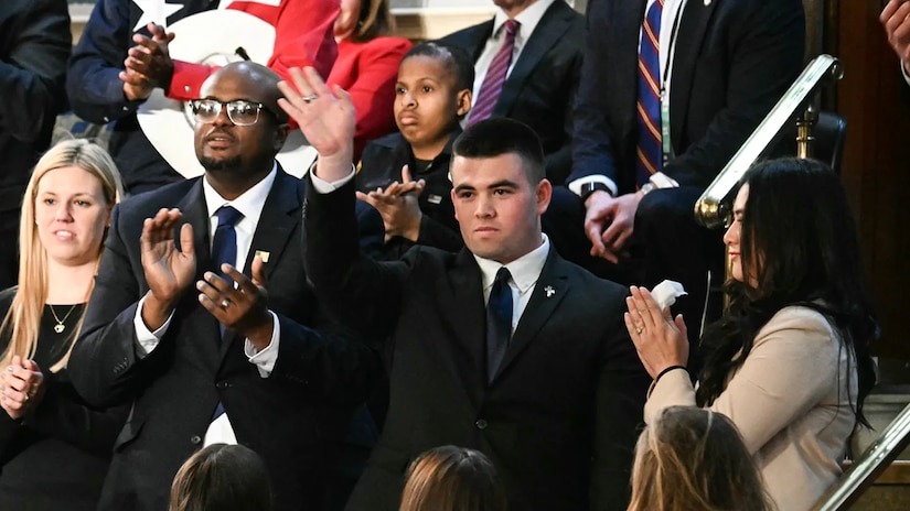 A young man wearing a suit waves to the crowd in an auditorium.