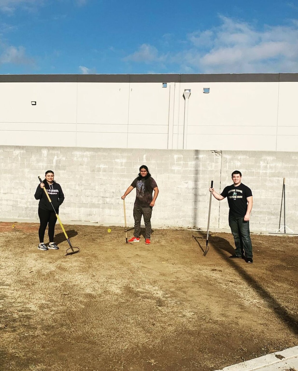 Three people stand on a dirt plain holding gardening rakes.