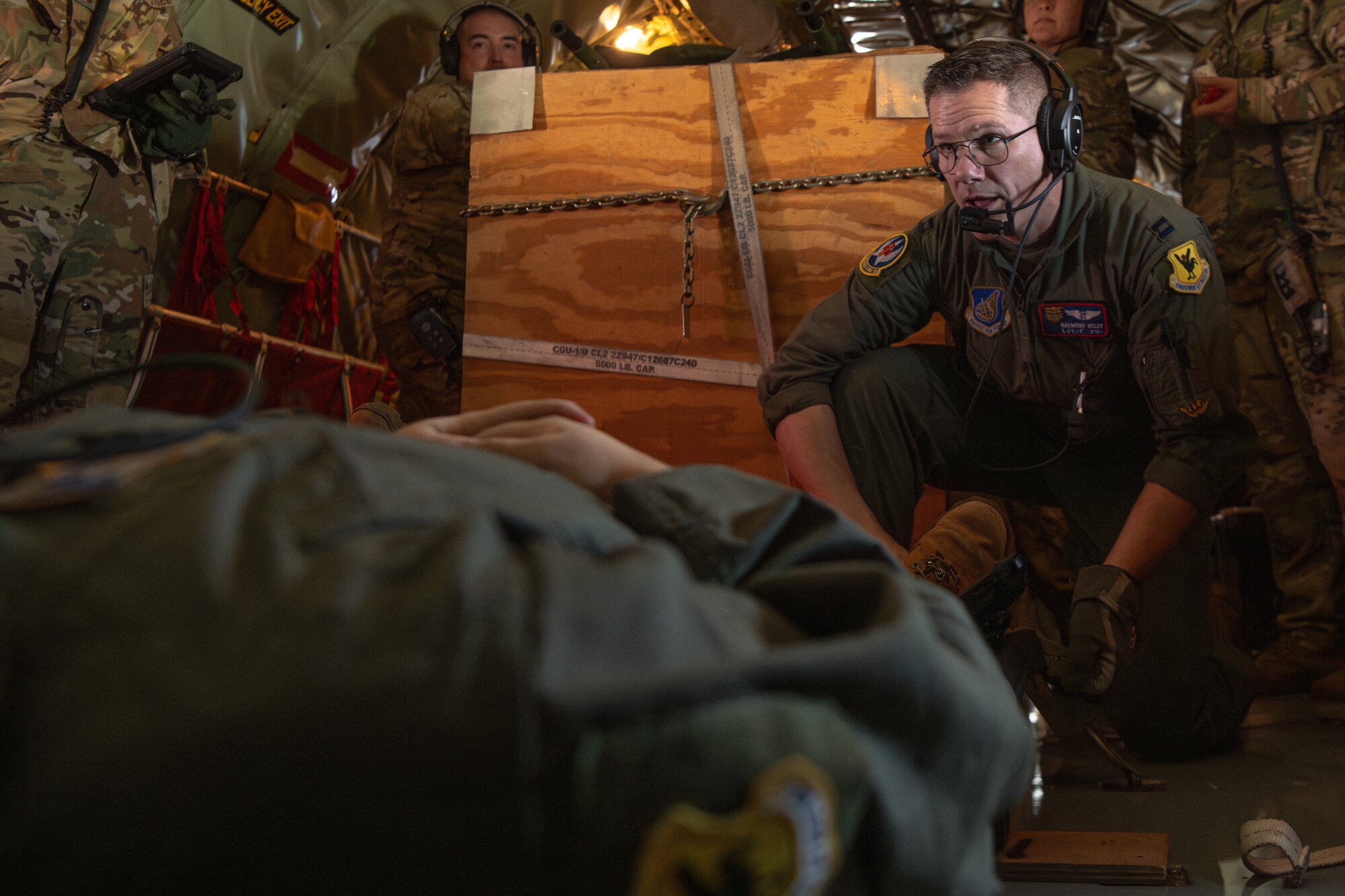 A man lays on a litter in the foreground while another man in the background secures the litter to the floor of the aircraft.