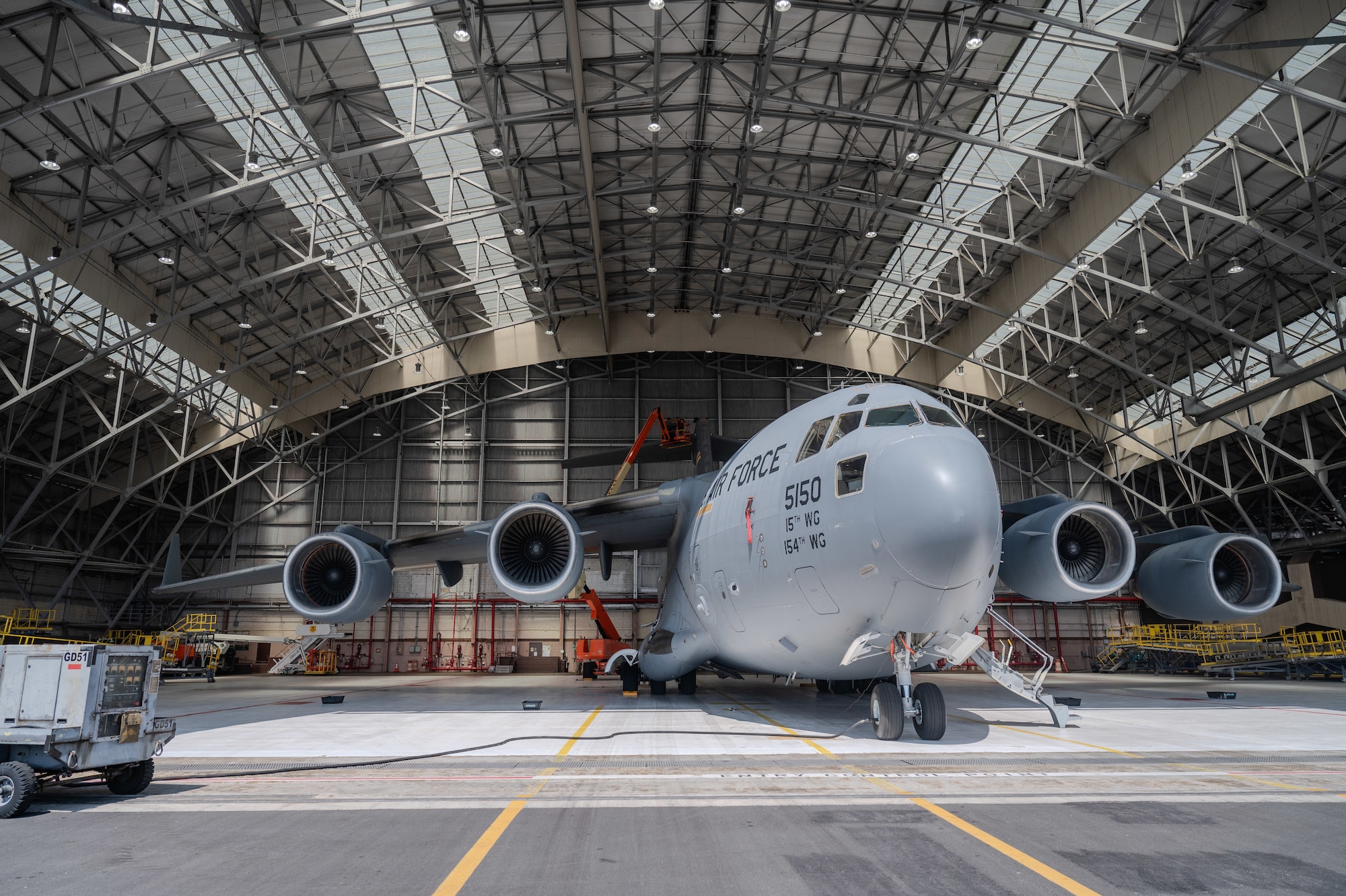 A C-17 Globemaster III aircraft sits inside a hangar. further away in the background several Airmen use a lift to