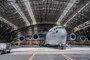 A C-17 Globemaster III aircraft sits inside a hangar. further away in the background several Airmen use a lift to