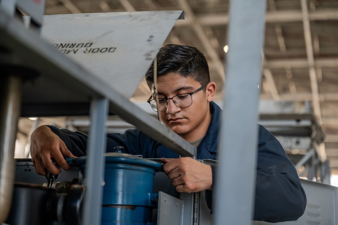 An Airman is wearing a blue suit and is attempting to use a small hand-held tool to unscrew a cap an oil filter.
