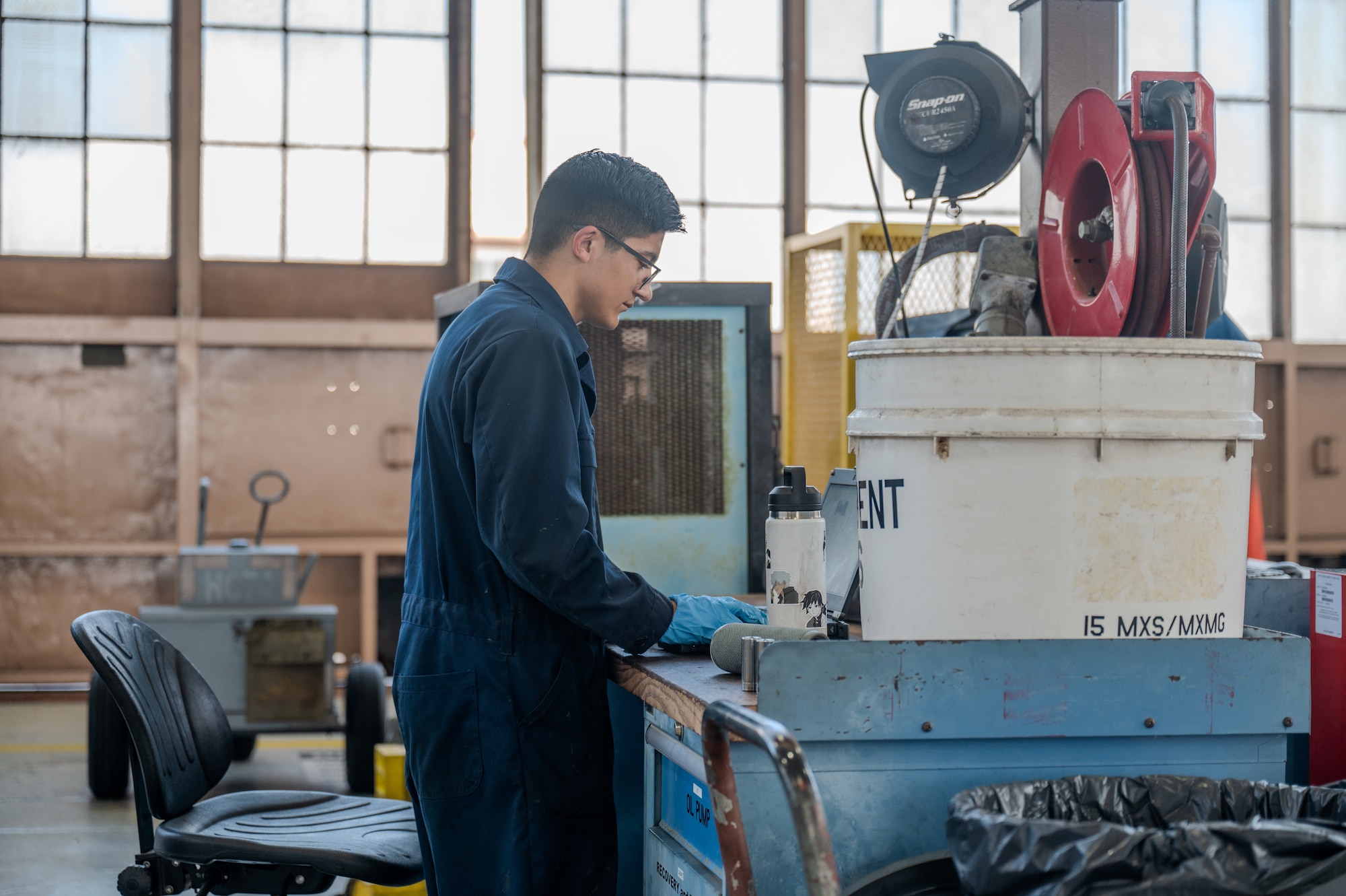 An airman uses a computer inside an aircraft hangar while wearing a blue work suit and gloves.