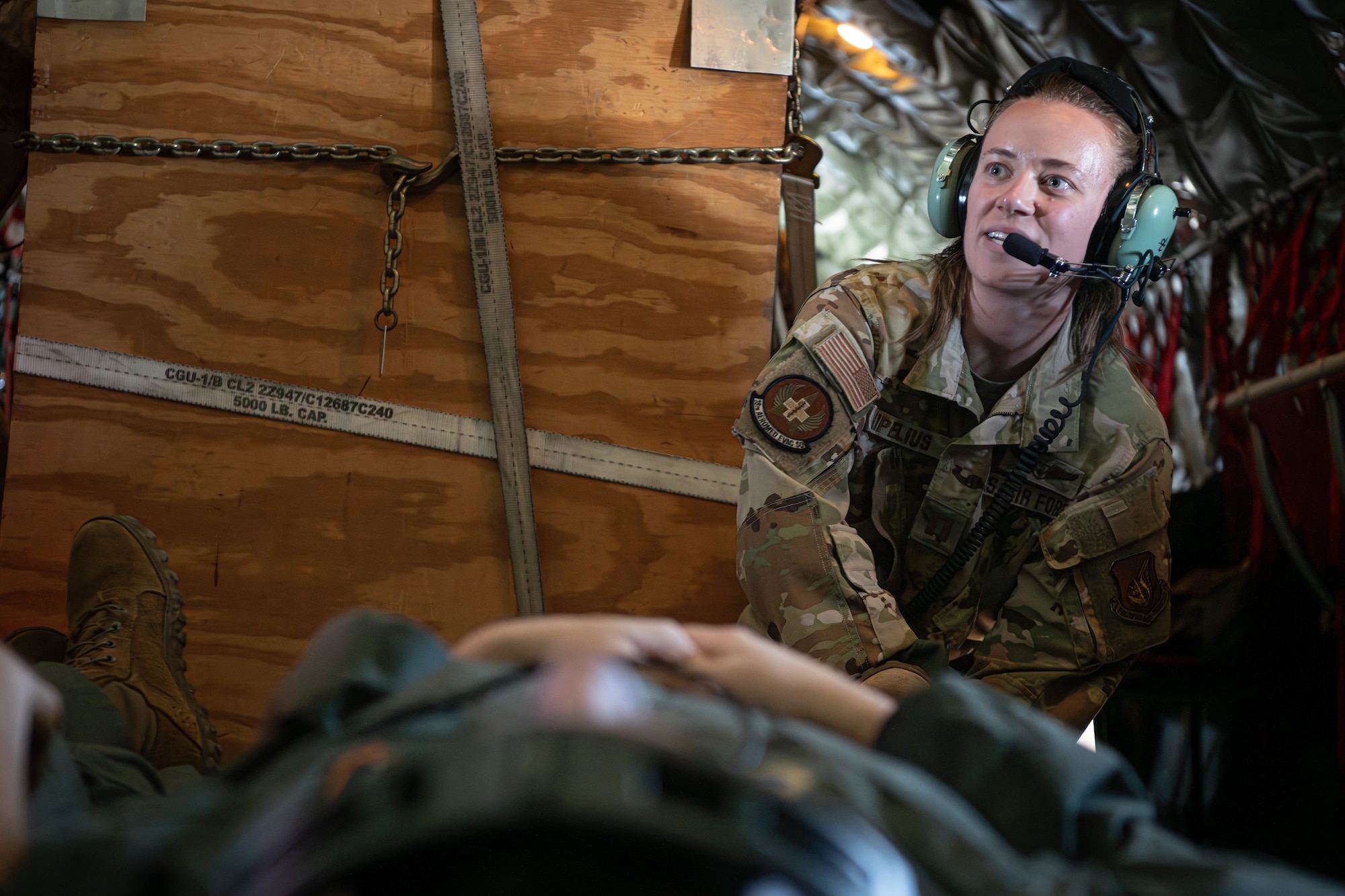 A man lays on a litter in the foreground while a woman in the background secures the litter to the floor of the aircraft.