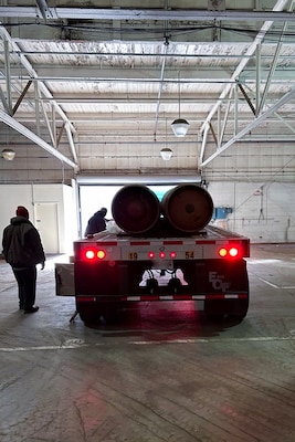 People in a large warehouse stand next to a flatbed truck that is loaded with red canisters.