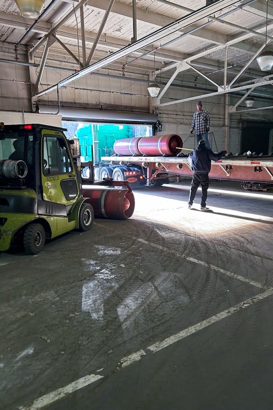 People in a large warehouse stand next to a flatbed truck that is loaded with red canisters.