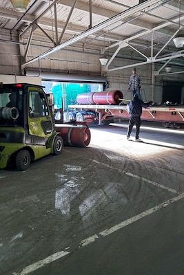 People in a large warehouse stand next to a flatbed truck that is loaded with red canisters.