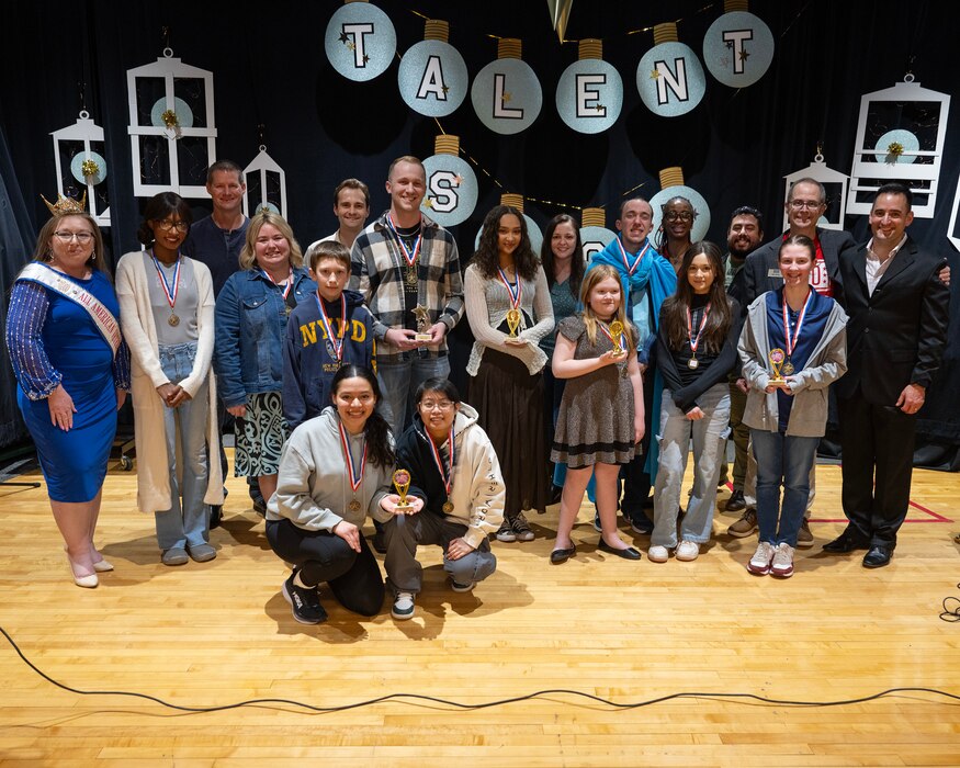 18 people pose for a group photo on a stage with a sign saying "Talent Show" behind them.