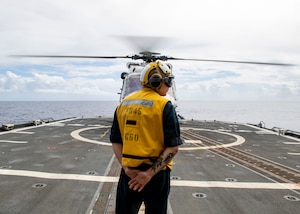 A man in a yellow vest and helmet on a flight deck of a ship signals to pilots in a helicopter preparing to take off.