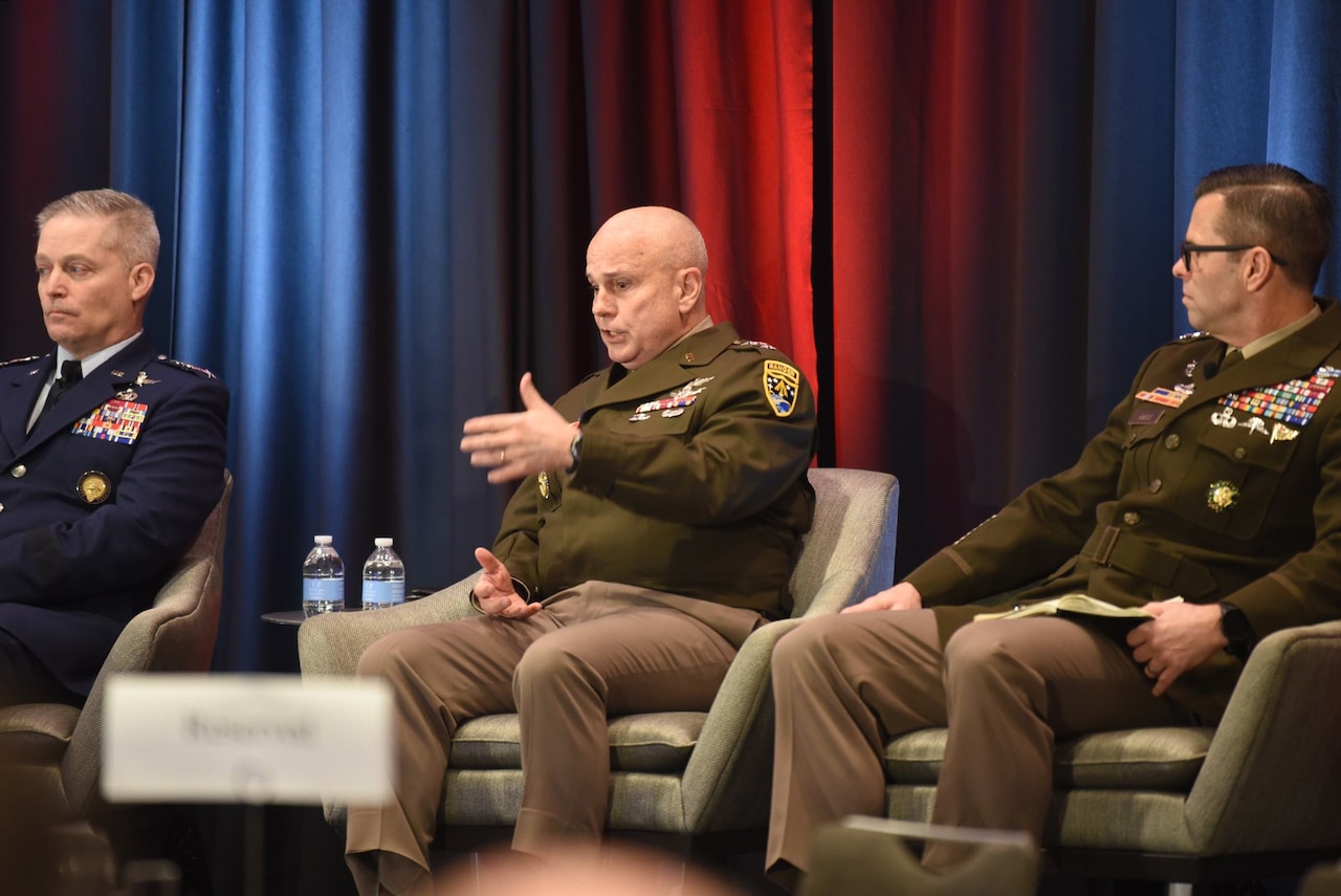 Three men dressed in military uniforms sit in chairs on a stage. The man in the middle is speaking.