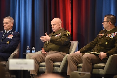 Three men dressed in military uniforms sit in chairs on a stage. The man in the middle is speaking.