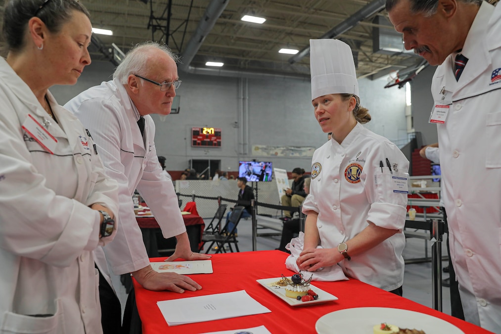 A chef speaks to three judges wearing white coats over a red table with a pastry on it.