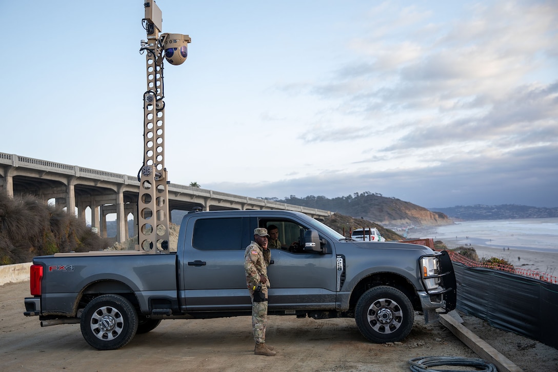 A soldier stands near a car near a bridge.