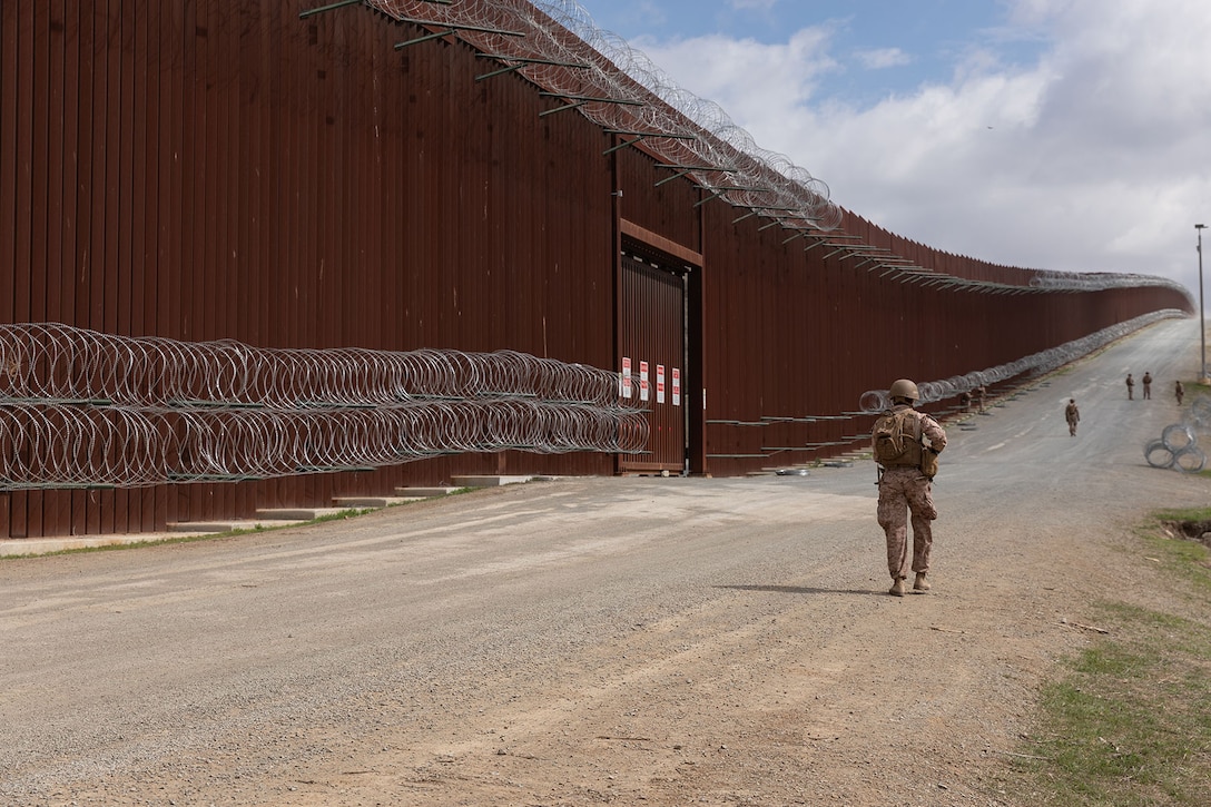 A Marine walks near a large fence under a cloudy, blue sky.