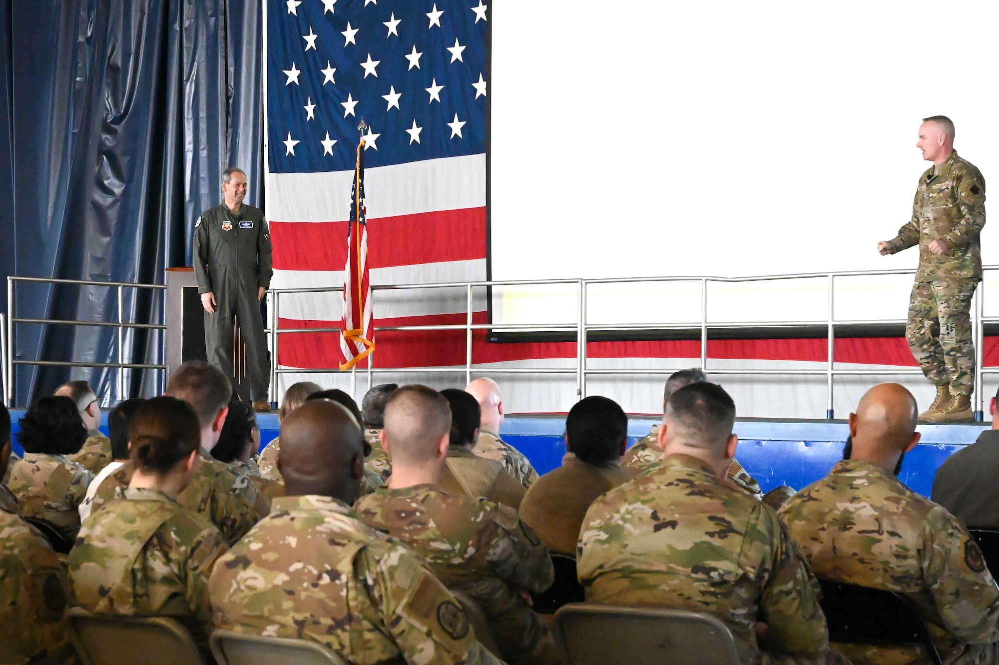 Group of Airman are getting briefed by two Airmen on stage.