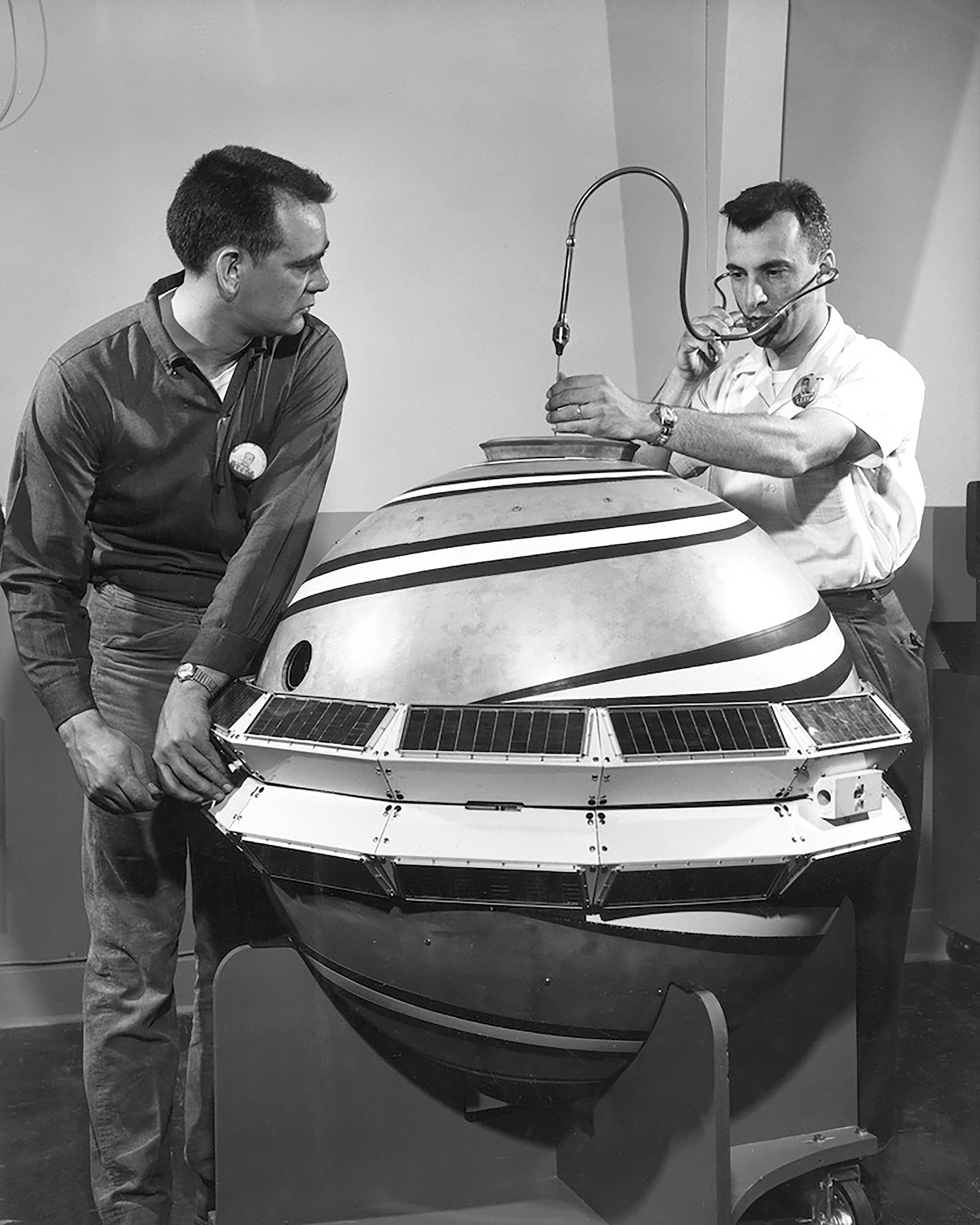 two men standing next to a round satellite. One man is holding what appears to be a stethoscope with one end in his ears and the other end he is holding at the top of the satellite.