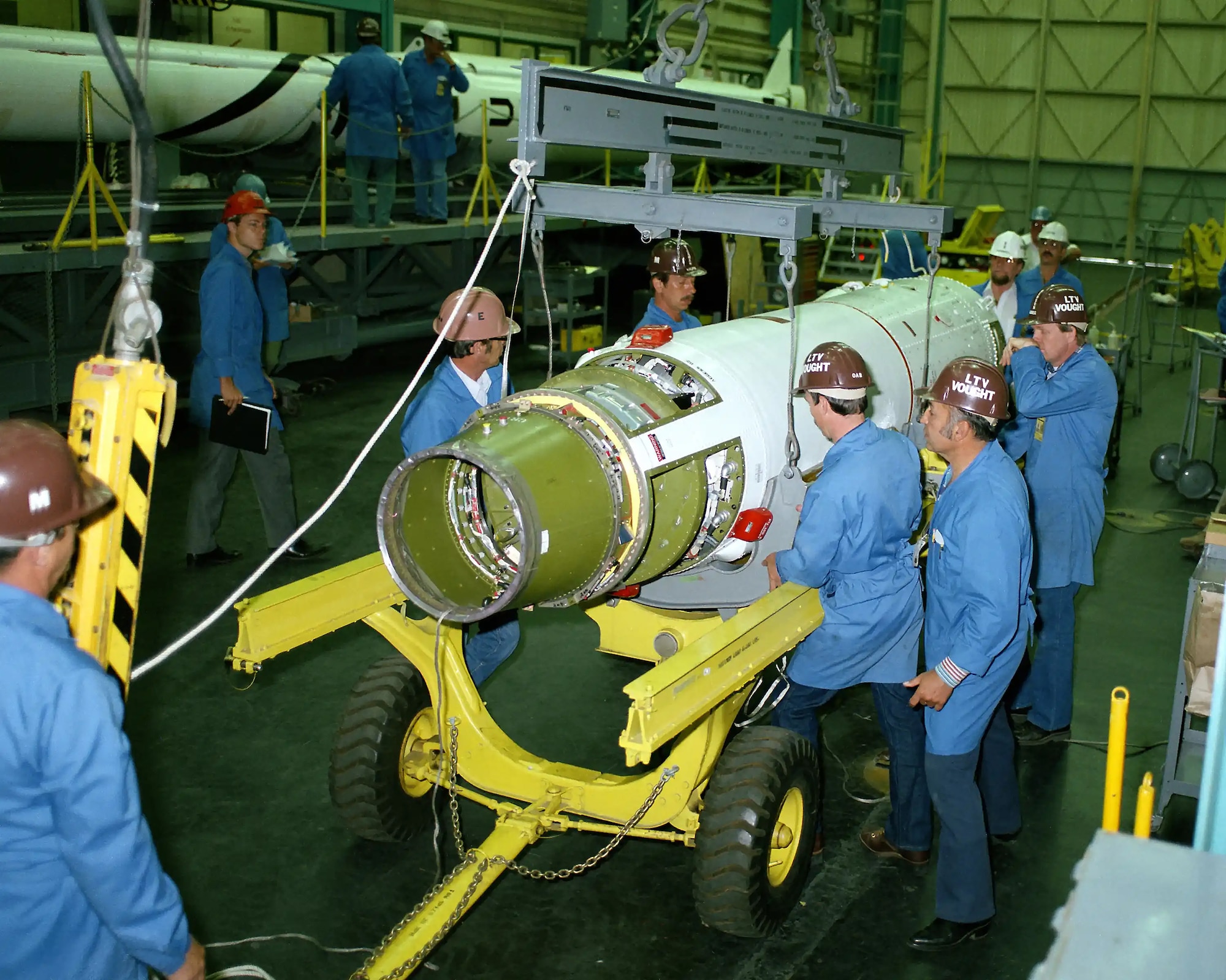 the Antares stage of the Scout rocket laying on its side on a yellow, wheeled cart surrounded by five men dressed in blue jumpsuits and wearing hardhats.
